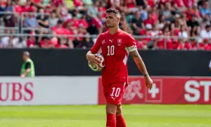Piłkarz w czerwonej koszulce z numerem 10 trzyma piłkę, na tle trybun i banerów.