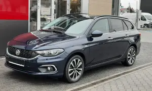 Ciemnoniebieskie kombi Fiat Tipo stoi przed budynkiem z czerwonym i szarym panelem.