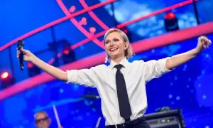Kasia Stankiewicz na scenie, w białej koszuli i krawacie, śpiewa z mikrofonem. W tle niebieska scenografia z czerwonymi elementami, przypominająca orła cień.