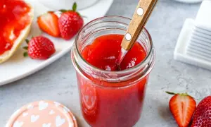 Ein Glas mit leuchtend roter Erdbeermarmelade, die mit einem Holzlöffel umgerührt wird. Daneben frische Erdbeeren und ein halbes Brötchen mit Marmelade – Erdbeermarmelade mit Pfiff für den perfekten Start in den Tag.