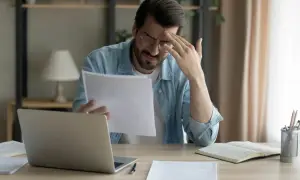 Mężczyzna zmartwiony przegląda dokumenty przy biurku z laptopem.