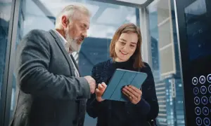 Starszy mężczyzna i młoda kobieta omawiają projekt na tablecie w windzie. To idealny moment na ich elevator pitch.