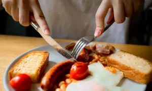Ręce krojące kiełbaskę na talerzu z fasolką, jajkiem sadzonym, pomidorkami i grzankami. To klasyczne angielskie śniadanie.