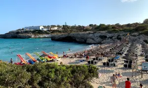 Słoneczna plaża Cala Domingos tętni życiem. Woda jest turkusowa, a na piasku widać rzędy parasoli i leżaków. Dzieci bawią się na kolorowych zjeżdżalniach.