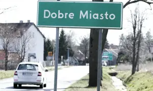 Zielony znak miejscowości "Dobre Miasto" wita podróżnych. W tle biały samochód i domy.