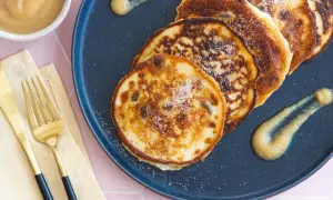 Goldbraune Quarkkeulchen ohne Kartoffeln, bestreut mit Zucker, auf einem dunklen Teller mit Apfelmus. Daneben liegen goldene Gabel und Messer.