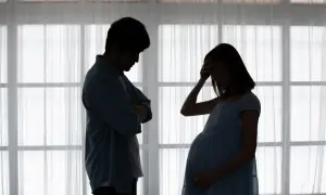 Ciężarna kobieta z ręką na czole i zmartwiony mężczyzna stoją naprzeciw siebie. Widać ich sylwetki na tle okna. To obraz strachu przed porodem.
