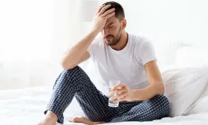 A man in pajamas sits on a bed, holding a glass of water and clutching his head, clearly sleeping off a hangover.