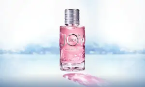 Różowy flakon Dior Joy 90ml z metalową nakrętką, na tle rozmytego, błękitnego krajobrazu.
