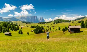 Ein Wanderer genießt die Aussicht auf eine Alm mit Berghütten und Wäldern. Perfekt für eine Mehrtageswanderung in den Dolomiten.