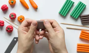 Ręce formują brązową masę, obok leżą warzywa z plasteliny i kolorowe paski do tworzenia animacji poklatkowej.