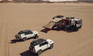 Un camion Dakar et deux 4x4 blancs dans le désert. L'équipe se réunit sous un auvent, prête pour l'aventure.