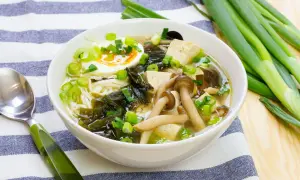 A miska zupy miso z tofu, jajkiem na pół, grzybami i szczypiorkiem. Obok leży zielona łyżka i pęczek dymki.