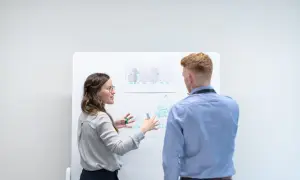Kobieta z okularami tłumaczy mężczyźnie schemat na tablicy, omawiając złożoność projektu i jego impedancję.