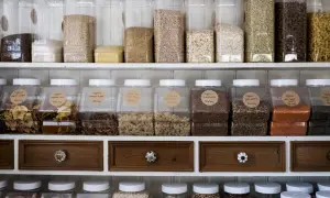 Regał do spiżarni pełen przezroczystych pojemników z różnymi produktami: makarony, kasze, płatki, suszone owoce i soczewica.