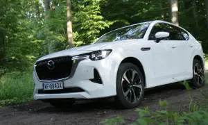 Biała Mazda CX-60 zaparkowana na leśnej ścieżce, otoczona zielenią drzew i traw.