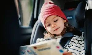 Kleines Mädchen mit roter Mütze liest ein Buch im Kindersitz. Perfekt für autofahrt spiele, um die Reise kurzweilig zu gestalten.