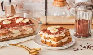 Ein köstliches Bananen Tiramisu, geschichtet mit Creme und Bananenscheiben, bestäubt mit Kakao. Daneben ein Glas Kakaopulver.