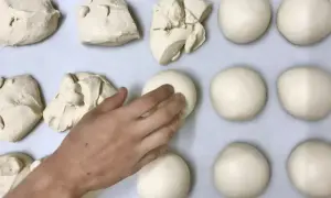 Des boules de pâte à pizza sont disposées sur une plaque. Une main façonne délicatement l'une d'elles.