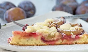 Ein saftiges Stück Apfel Pflaumen Kuchen mit Streuseln, bestäubt mit Puderzucker. Im Hintergrund sind frische Pflaumen zu sehen.