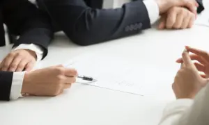 Osoby w garniturach analizują dokumenty, w tym poświadczenie dziedziczenia. Jedna z nich wskazuje piórem na tekst.