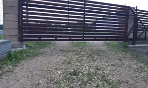 Brązowa, metalowa brama przesuwna z poziomymi szczeblinami. Widoczna jest szczelina pod bramą, przez którą widać trawę i żwir.