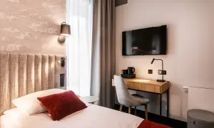 Przytulny pokój w hotelu Vesper House: łóżko z czerwonymi akcentami, biurko z czajnikiem i telewizorem, szare zasłony.