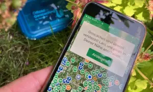 Smartfon pokazuje mapę z ukrytymi skarbami. To właśnie geocaching co to za zabawa! W tle niebieskie pudełko z napisem "Official Geocaching".