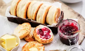 En korg med gyllene engelska muffins, en burk sylt och smör. En muffin är delad och bredd med smör och sylt.