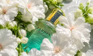 Szklana butelka perfum Jardin Bohème Escapade Aquatique, otoczona białymi kwiatami. Ten zapach to jak szczęśliwy los na lato.