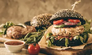 Gotowy hamburger z czarną bułką, kotletem wołowym i warzywami, obok surowy kotlet i sos.