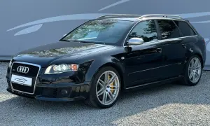 Czarny Audi RS4 B7 Avant z żółtymi zaciskami hamulcowymi, zaparkowany na żwirze przed szarą ścianą z grafiką.
