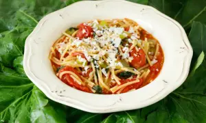 Un piatto di pasta con i tenerumi asciutta, condita con pomodoro fresco e formaggio grattugiato, adagiato su foglie verdi.