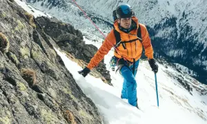 Alpinista w pomarańczowej kurtce wspina się po skalistym, ośnieżonym zboczu, używając czekana alpinisty inaczej jako wsparcia.