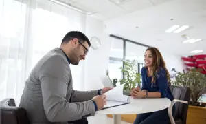 Uomo in giacca grigia firma documenti, donna sorride. Potrebbe essere un colloquio o una discussione su un nuovo progetto di job rotation.