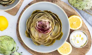 Ein gekochter Artischocke auf einem Teller, bereit für ein leckeres Artischocken Rezept. Dazu Zitronenspalten und eine Dip-Schale.