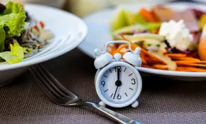 Réveil miniature sur une table, symbolisant le jeûne intermittent 12h. Des assiettes de salade et de légumes sont floues en arrière-plan.