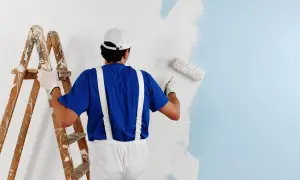 Malarz w niebieskiej koszulce i białych ogrodniczkach maluje ścianę na jasnoniebieski kolor. Obok stoi drewniana drabina. Czas schnięcia farby zależy od wielu czynników.