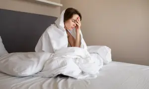 Kobieta w łóżku, przykryta pościelą, z ręką na czole. Wygląda na zmęczoną i zmartwioną, być może przez koszmary w ciąży.