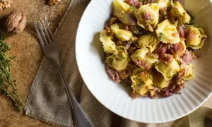 En skål med tortellini prosciutto, valnötter och timjan. En gaffel ligger bredvid på en rustik träyta.