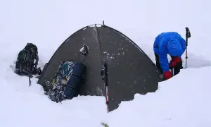 Osoba w niebieskiej kurtce i czerwonych rękawiczkach przy namiocie w śniegu. Obok plecaki i kijki, jak złożyć kijki do nordic walking.