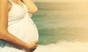 Kobieta w białej sukience na plaży, z dłonią na brzuchu. Czy w ciąży można chodzić na solarium? Słońce na plaży jest naturalne.