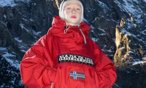 Modelka w czerwonej kurtce z logo Napapijri Geographic i flagą Norwegii na tle ośnieżonych gór. Napapijri co to za firma? To marka odzieży outdoorowej.