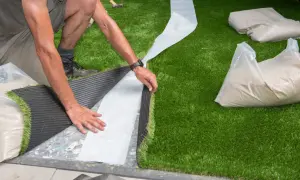 Mann verlegt Kunstrasen, legt die Nahtstelle frei und bereitet sie für das Verkleben vor. Sand gefüllt Säcke liegen bereit.