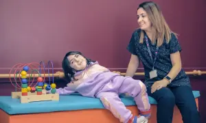 Uśmiechnięta dziewczynka w fioletowym dresie leży obok terapeuty, bawiąc się zabawką z koralikami.