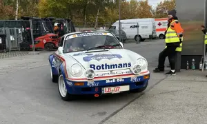 Ein weißer Porsche mit Rothmans-Aufschrift und dem Kennzeichen LB 07. Im Hintergrund stehen Gabelstapler und ein Rettungswagen.