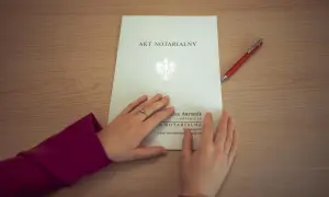 Dłonie na akcie notarialnym, obok czerwony długopis. Dokument zawiera pieczęć państwową i dane notariusza.