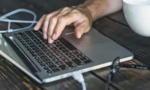 Dłoń na klawiaturze laptopa, biały kabel podłączony. Czas dowiedzieć się, jak zresetować laptopa.