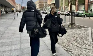 Dwie osoby w czarnych kurtkach puchowych idą chodnikiem, niosąc torby. Proste, miejskie kurtka puchowa stylizacje.