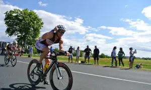 Triathlonista w kasku i okularach ściga się na rowerze, a widzowie dopingują go na poboczu. To część kalendarza triathlonowego.